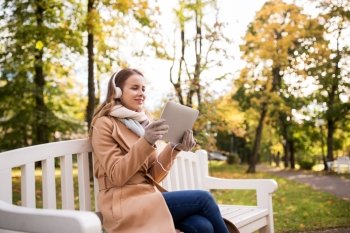 season, technology and people concept - beautiful happy young woman with tablet pc computer and headphones listening to music and sitting on bench in autumn park
