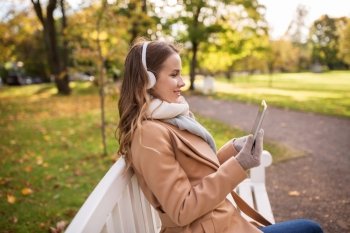 season, technology and people concept - beautiful happy young woman with tablet pc computer and headphones listening to music and sitting on bench in autumn park