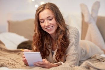 christmas, winter, holidays and people concept - happy young woman with pencil and notebook writing in bed at home bedroom