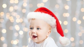 childhood, christmas, holidays and people concept - beautiful little baby boy in christmas santa hat over blue lights background