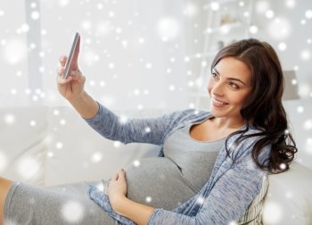 pregnancy, winter, technology, people and expectation concept - happy pregnant woman with smartphone taking selfie at home over snow