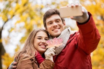 love, technology, relationship, family and people concept - smiling couple with maple leaf taking selfie by smartphone in autumn park