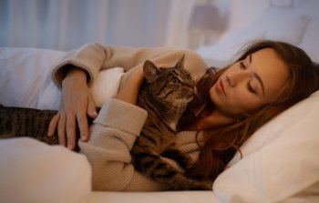 pets, comfort, rest and people concept - happy young woman with cat lying in bed at home at night