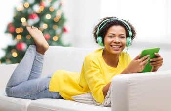 people, technology, holidays and leisure concept - happy african american young woman lying on sofa with tablet pc computer and headphones listening music at home over christmas tree lights background