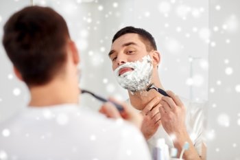 beauty, hygiene, shaving, grooming and people concept - young man looking to mirror and shaving beard with manual razor blade at home bathroom over snow