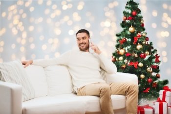 technology, people and holidays concept - smiling man calling on smartphone over christmas tree and lights background