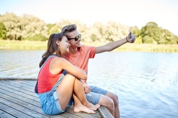 holidays, vacation, love and people concept - happy teenage couple taking selfie on smartphone and sitting on river berth at summer