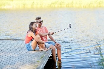 holidays, vacation, love and people concept - happy teenage couple taking picture by smartphone selfie stick and sitting on river berth at summer