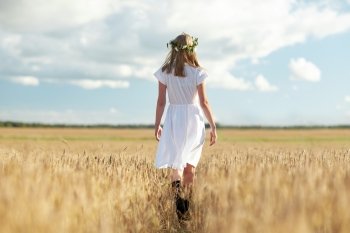 happiness, nature, summer holidays, vacation and people concept - smiling young woman in wreath of flowers and white dress walking along cereal field