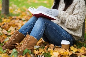 season, literature, education and people concept - close up of young woman reading book and drinking coffee from paper cup in autumn park