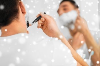 beauty, shaving, grooming and people concept - close up of young man looking to mirror and shaving beard with manual razor blade at home bathroom over snow