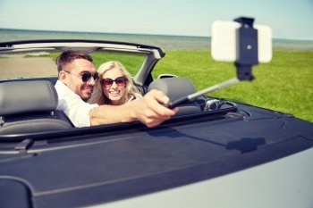 road trip, travel, couple, technology and people concept - happy man and woman driving in cabriolet car and taking picture with smartphone on selfie stick