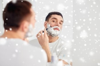 beauty, grooming, winter, christmas and people concept - young man looking to mirror and shaving beard with manual razor blade at home bathroom over snow