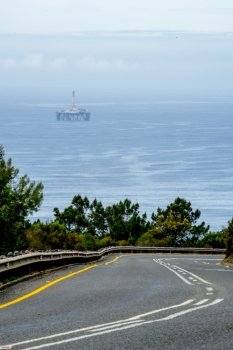An open road at a mountain pass lies in front of the ocean with an oil drill platform in the far distance.