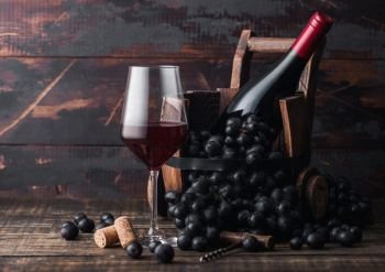 Elegant glass of red wine with dark grapes and bottle of wine inside vintage wooden barrel on dark wooden background. Wine still life concept.