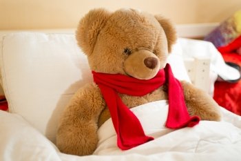 Closeup photo of brown teddy bear in red scarf lying in bed