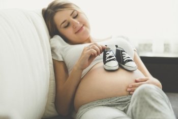 Toned photo of happy pregnant woman lying on sofa and holding baby boots