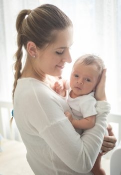 Closeup portrait of beautiful young mother holding newborn baby at big window