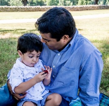 Latino father sitting outside on the grass holding his son who is playing with a toy car