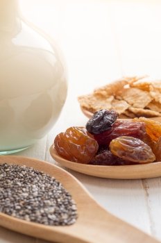 Set of raisins, whole wheat grain flakes and chia seeds in wooden spoon and milk on white wood background with selective focus point
