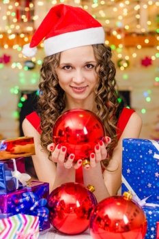 Young beautiful girl sitting at a table with Christmas gifts in the Christmas atmosphere. The girl is holding a big Christmas ball