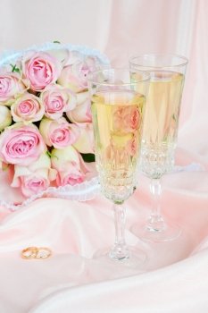 Bouquet of pink roses, wedding rings and two glasses of white wine on satin fabric background