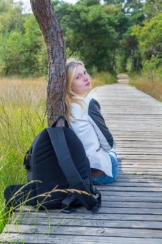 Blonde dutch teenage girl sitting on wooden path in nature with backpack