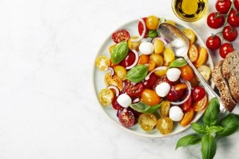 Blue plate of fresh and healthy Mediterranean salad with mozzarella cheese, tomatoes, red onion, basil leaves and a spoon on white marble vintage background.