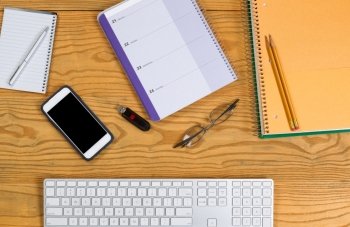 High angled view of desktop consisting of computer keyboard, pencils, pen, calendar, reading glasses, cell phone, notepad and thumb drive. Horizontal layout.
