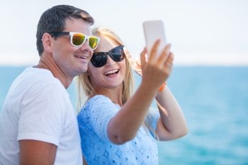 Happy young man and woman in fashionable sunglasses taking cellphone selfie on background of defocused blue sea. Vacation photos. Happy summer selfie of young couple in sunglasses