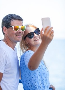 Smiling couple in sunglasses making happy summer selfie with cell against blue sea and sky background. Cheerful couple in sunglasses taking mobile selfie