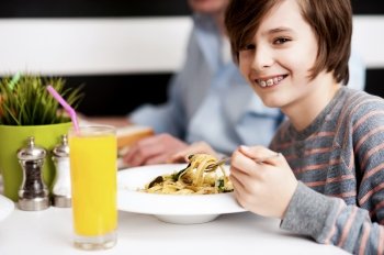 Boy in a striped t-shirt eating pasta with glass of fresh orange juice on table