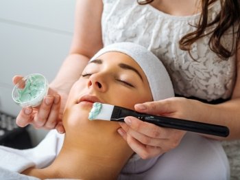 Photo of a young beautiful girl receiving a green facial mask in spa beauty salon.
