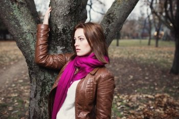 Pensive young  brunette girl standing near tree in autumn park