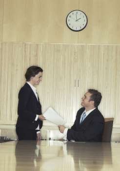 Business woman hands over papers to business man who is sitting at the head of the conference table