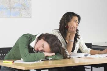 Two teenage girls in class, one has fallen asleep and the other is looking bored out of the window