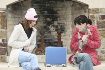 Two beautiful young woman (hispanic and caucasian) sitting outside by the fireplace with laptop. Wearing winter clothes.