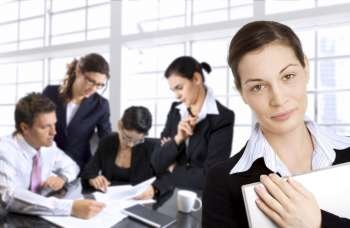 Young businesswoman looks at the camera while her team is brainstorming in the background.