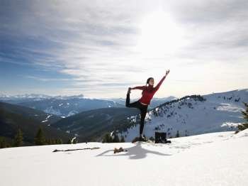 Young woman doing yoga pose on rock in snow in mountainous terrain.