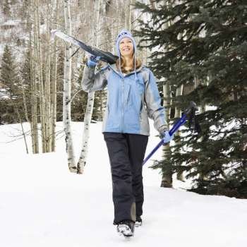 Mid adult Caucasian female skier wearing blue ski clothing walking and carrying skis on shoulder.
