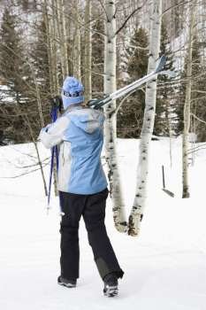 Rear view of mid adult Caucasian female skier wearing blue ski clothing walking and carrying skis on shoulder.