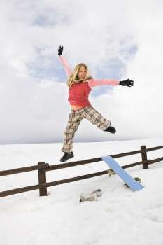 Young woman in winter clothes by fence in snowy field with snowboard jumping into air excitedly.