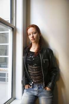 Pretty redhead young woman standing indoors by window leaning against wall with hands in jean pockets.
