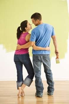 Attractive couple standing in front of partially painted wall with arms around eachother smiling.
