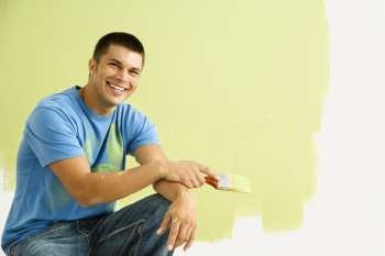 Smiling man kneeling in front of partially painted wall holding paintbrush.