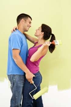 Couple hugging and smiling at eachother holding paintbrushes in front of partially painted wall.