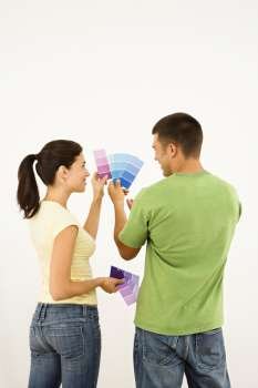 Attractive young adult couple holding color paint samples up to white wall.