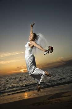 Young adult female Caucasian bride jumping on beach.