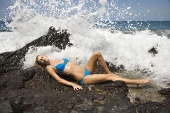Young adult female Caucasian lying on rock in bikini with wave crashing.