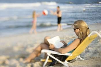 Caucasian mother reading in lounge chair on beach while husband and daughter play ball in background.
